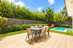 Sunlit backyard patio with a modern dining set overlooking a small pool, surrounded by lush greenery and a privacy fence. Clean lines, warm wood tones, and bright natural light create a calm, inviting outdoor space.