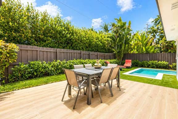 Sunlit backyard patio with a modern dining set overlooking a small pool, surrounded by lush greenery and a privacy fence. Clean lines, warm wood tones, and bright natural light create a calm, inviting outdoor space.